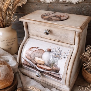 Puede incluir: Una panera de madera con acabado marrón claro, que cuenta con una puerta abatible y un cajón superior. La puerta muestra una ilustración impresa de pan, una botella de vidrio y flores. Tiene dos pequeños pomos de cerámica con diseño azul y blanco.