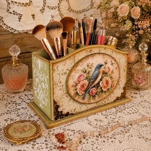 Puede incluir: Un organizador de maquillaje rectangular de madera verde salvia con un panel ovalado central que muestra una ilustración de un pájaro azul y rosas rosadas. El organizador tiene varios compartimentos para brochas, labiales y rímel.