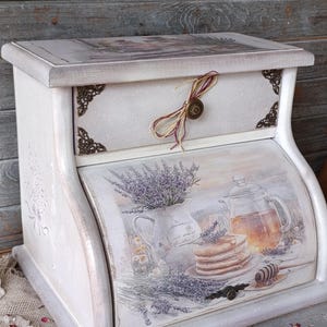 Könnte beinhalten: Eine weiße Brotdose aus Holz mit einer Schublade und einer gewölbten Front. Die Box ist mit floralen und Lebensmittelbildern dekoriert, darunter Lavendel, Pfannkuchen und Honig. Sie hat dekorative Metallecken und einen Knopfverschluss.