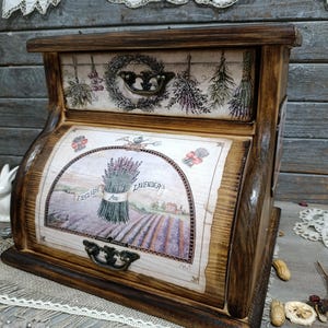 Puede incluir: Una caja de almacenamiento de madera con dos cajones, con un diseño rústico. Los cajones están adornados con imágenes de lavanda y hierbas, con la palabra "LAVENDER" visible. La caja tiene un acabado marrón oscuro y tiradores de metal ornamentados.