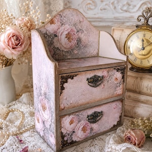 May include: A wooden desktop organizer featuring two drawers and an open top shelf. The exterior is decorated with a soft pink and grey floral pattern, vintage script text, and ornate dark metal drawer pulls with matching corner accents.