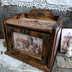 Puede incluir: Una panera de madera con acabado marrón oscuro. La parte delantera presenta una imagen enmarcada de productos horneados y artículos de cocina. Los detalles incluyen esquinas decorativas de metal y un asa. La parte superior tiene un diseño impreso de productos horneados.