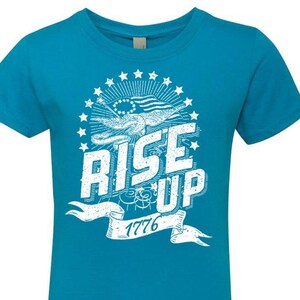 Peut inclure: Un t-shirt bleu turquoise avec un motif blanc repr&eacute;sentant un aigle, des &eacute;toiles et le texte "Rise Up 1776".