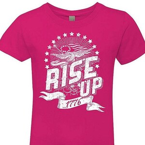 Peut inclure: Un t-shirt rose avec un motif graphique blanc repr&eacute;sentant un aigle vieilli tenant une banni&egrave;re sur laquelle est inscrit "Rise Up 1776". L'aigle tient un drapeau &agrave; &eacute;toiles et &agrave; rayures. Le motif est entour&eacute; d'un cercle d'&eacute;toiles.