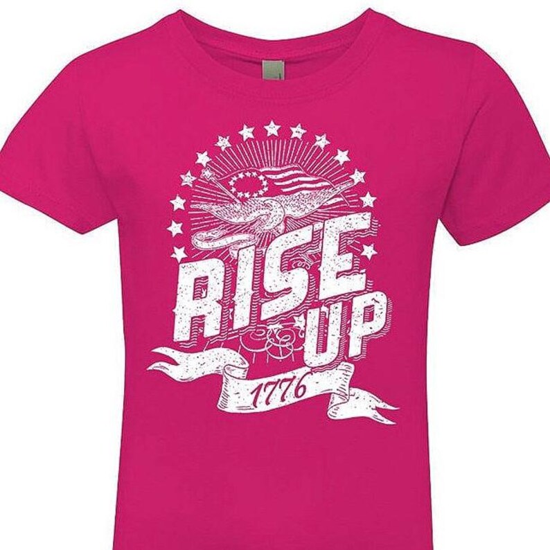 Peut inclure: Un t-shirt rose avec un motif graphique blanc repr&eacute;sentant un aigle vieilli tenant une banni&egrave;re sur laquelle est inscrit "Rise Up 1776". L'aigle tient un drapeau &agrave; &eacute;toiles et &agrave; rayures. Le motif est entour&eacute; d'un cercle d'&eacute;toiles.