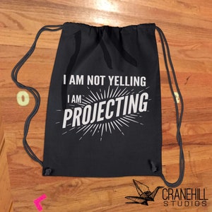 May include: Black drawstring backpack with white text that reads "I AM NOT YELLING I AM PROJECTING". The text is in a bold, sans-serif font. The backpack has black drawstrings and is made of a soft material.