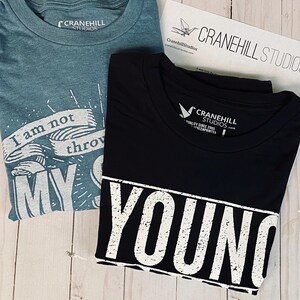 May include: Two t-shirts, one teal blue and one black. The teal blue t-shirt has the text "I am not throwing my shot" printed on it. The black t-shirt has the text "Young and Reckless" printed on it in white.