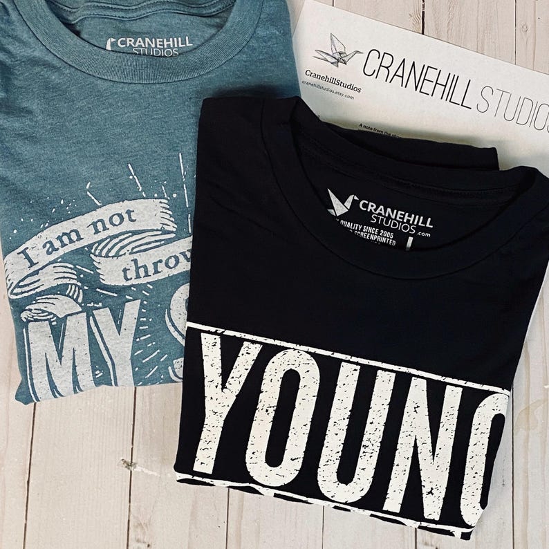 May include: Two t-shirts, one teal blue and one black. The teal blue t-shirt has the text "I am not throwing my shot" printed on it. The black t-shirt has the text "Young and Reckless" printed on it in white.