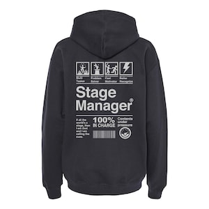 May include: A dark gray hooded sweatshirt with a white graphic print that reads "Stage Manager" and includes a list of skills and responsibilities. The text "100% In Charge" and "Contents under pressure" are also included in the graphic.