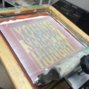 May include: A close-up of a screen printing frame with a design that reads "Young Scrappy Hungry" in yellow and green lettering on a red background. The design is being prepared for printing.