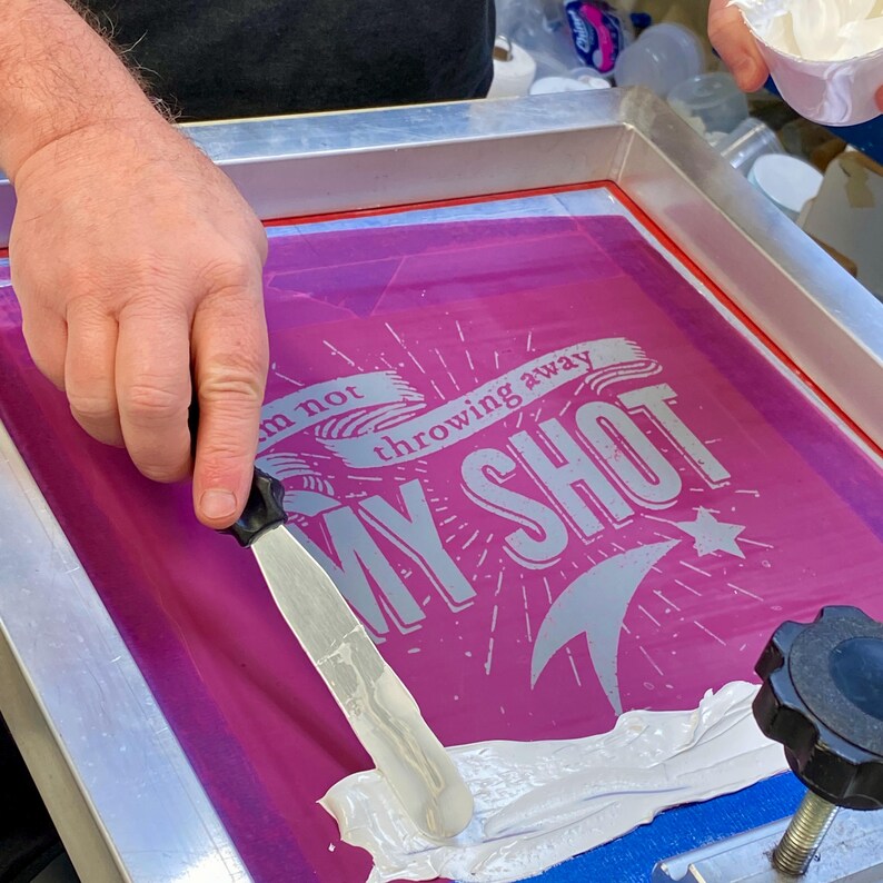 Peut inclure: Une personne utilise une raclette pour &eacute;taler de l'encre blanche sur un cadre de s&eacute;rigraphie. Le cadre est rose et porte l'inscription "I'm not throwing away my shot".