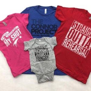 May include: Four t-shirts in different colours and designs. A pink t-shirt with the text "I am not throwing away my shot" and a star. A blue t-shirt with the text "The Connor Project You Will Be Found". A red t-shirt with the text "Straight Outta Rehearsal". A grey baby onesie with the text "Young Scrappy Hungry".