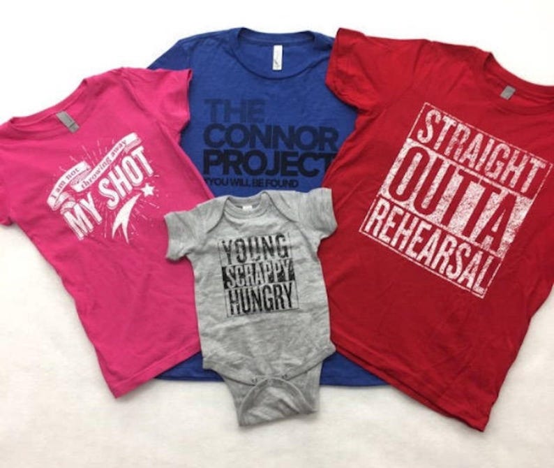 May include: Four t-shirts in different colours and designs. A pink t-shirt with the text "I am not throwing away my shot" and a star. A blue t-shirt with the text "The Connor Project You Will Be Found". A red t-shirt with the text "Straight Outta Rehearsal". A grey baby onesie with the text "Young Scrappy Hungry".