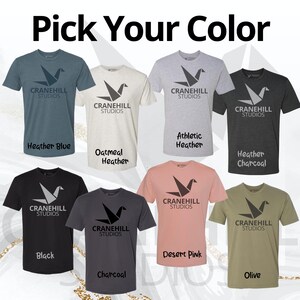 May include: A selection of t-shirts in various colours, each featuring a grey origami crane logo with the text "Cranehill Studios". The colours include heather blue, oatmeal heather, athletic heather, heather charcoal, black, desert pink, charcoal, and olive.