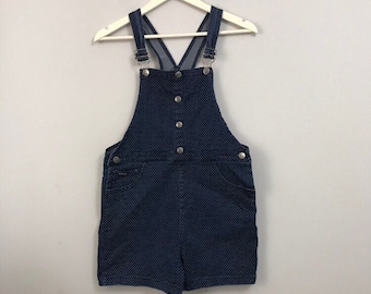 May include: A dark blue denim overall dress with white polka dots and a button closure. The dress has adjustable straps and pockets.