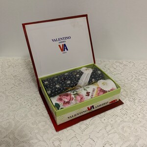 May include: A red box with a green lid containing two handkerchiefs. The handkerchiefs are wrapped in tissue paper and have floral patterns. The box has the text "VALENTINO AMEDEO VA ITALIA" on the front.