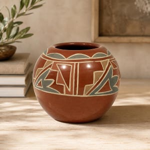 May include: A round, terracotta-colored ceramic vase featuring a wide opening and a smooth, polished finish. The exterior is decorated with a repeating geometric pattern in muted sage green, tan, and white lines encircling the upper body.
