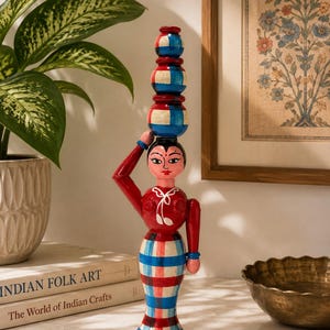 May include: A hand-painted wooden figurine of a person with a red top and a blue, red, and white plaid skirt. The figure has one arm raised to balance a vertical stack of three small, matching plaid pots on their head, featuring painted facial details.
