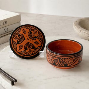 May include: A round terracotta lidded bowl featuring a vibrant orange base color with intricate black hand-painted floral and geometric patterns. The circular lid is displayed upright next to the open bowl, both showcasing matching decorative motifs.