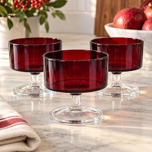 May include: Three matching dessert bowls made of deep red glass. Each bowl features a wide, cylindrical basin supported by a short, clear glass stem and a circular, flat base. The bowls are arranged in a cluster on a light-colored surface.