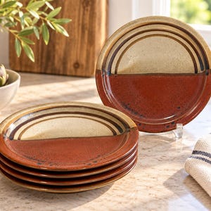May include: A set of five round ceramic plates featuring a two-tone design with a speckled cream upper half and a solid terracotta lower half. The plates are decorated with dark brown concentric circular bands separating the two distinct color sections.