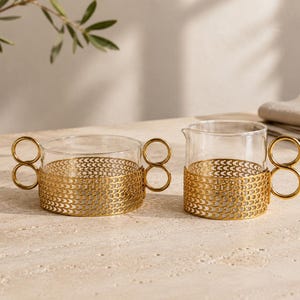 Pode incluir: Um açucareiro de vidro transparente e uma leiteira a condizer, cada um inserido numa manga decorativa de metal dourado com um padrão de chevron repetitivo e com pegas distintas de duplo anel nas laterais dos recipientes de vidro.