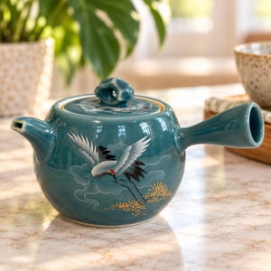May include: A teal ceramic teapot with a side handle and a lid featuring a knob. The body is decorated with a painted white and black crane in flight, surrounded by stylized clouds and gold-colored pine needles on a smooth, glossy surface.