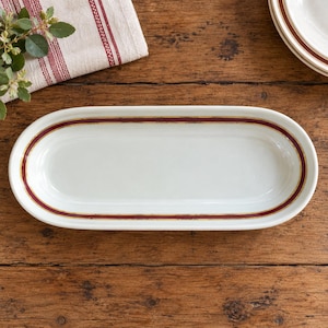 May include: An oval-shaped white ceramic serving dish featuring a decorative border consisting of a deep red band framed by thin gold lines. The dish has a smooth, glossy finish and a shallow basin, centered horizontally on a dark wooden surface.