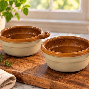 May include: Two round ceramic bowls with a single small handle on each side. The bowls feature a smooth, light cream-colored exterior and a contrasting speckled, glossy brown interior and rim, designed for serving individual portions of food.