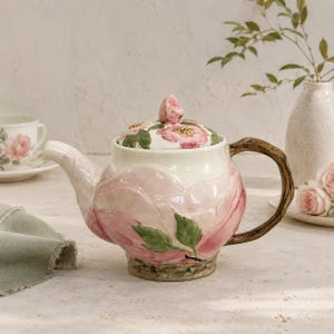 Puede incluir: Una tetera de cerámica con un diseño floral rosa esculpido en el cuerpo y una tapa a juego adornada con flores rosas pintadas. El asa tiene una textura que imita una rama de árbol marrón y la base presenta un acabado rústico.