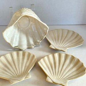 Könnte beinhalten: Ein Set aus vier cremeweißen Keramikschalen in Form von Jakobsmuscheln. Eine Schale steht aufrecht auf einem transparenten Ständer, während die anderen drei flach liegen. Jedes Stück hat eine glänzende Oberfläche mit feinen beigen Rillen.