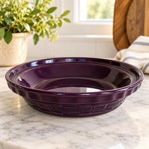 May include: A deep purple ceramic pie dish with a glossy finish and a textured exterior featuring a woven basket-weave pattern. The round baking dish has a wide, smooth rim and a slightly flared edge, designed for baking pies or tarts.