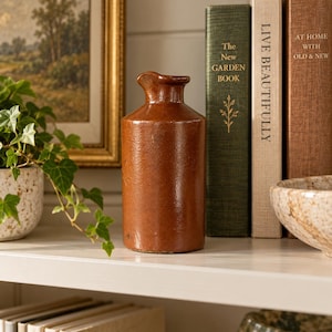 May include: A cylindrical brown stoneware bottle with a smooth, glossy finish and a small, slightly flared spout at the top. The bottle features a simple, uniform design with a flat base and a narrow neck, standing upright as a decorative object.