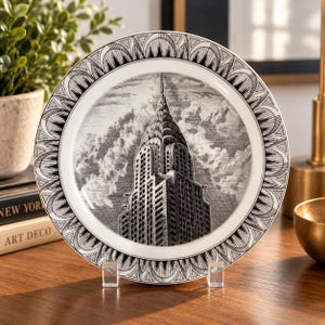 May include: A round ceramic plate featuring a black and white illustration of the Chrysler Building in New York City. The plate has a decorative border with a repeating arched pattern and is displayed on a clear acrylic stand.