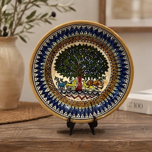 May include: A round ceramic decorative plate featuring a central hand-painted scene of a large tree surrounded by various animals including a lion and deer. The plate has a wide border with intricate blue, brown, and white geometric patterns.