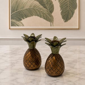 May include: Two pineapple-shaped candle holders made of metal with a bronze-toned body and a green-toned leafy top. Each piece features a textured, cross-hatched pattern on the base and a circular opening at the top for holding a candle.