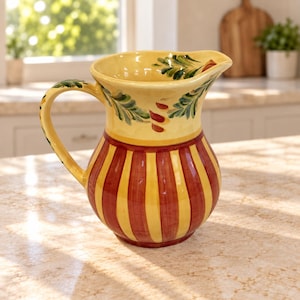 May include: A ceramic pitcher featuring a yellow base with vertical red stripes on the body. The upper section is yellow with a hand-painted green leaf and red accent pattern around the rim. It includes a curved handle and a pouring spout.
