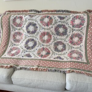 May include: A woven throw blanket featuring a repeating pattern of floral wreaths in shades of pink, purple, and green on a white background. The blanket has a decorative border with a pink and white lattice design and a fringed edge on all sides.