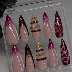 May include: A set of ten stiletto-shaped press-on nails in a clear plastic case. The designs include a nude base with deep magenta French tips, gold studs, horizontal dark bands, gold chains, and a dark magenta animal print pattern on the nails.