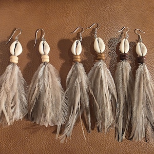 May include: Three pairs of dangle earrings featuring silver-toned hooks, white cowrie shells, and small wooden beads above long, grey, natural-looking feathers. The earrings are arranged in a row, showcasing the consistent design of the shells and feathers.