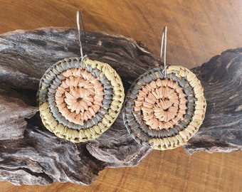 May include: A pair of circular drop earrings featuring concentric rings of woven raffia in shades of peach, charcoal gray, and light tan. Each earring is attached to a thin, silver-toned metal hook wire for suspension from the earlobe.
