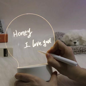 May include: A white light-up bulb shaped sign with a clear writing surface. The sign is lit up and has the words "Honey I love you" written on it in white marker.
