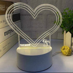 May include: A white heart-shaped acrylic night light with a small cat silhouette on the bottom. The light is on a white circular base.