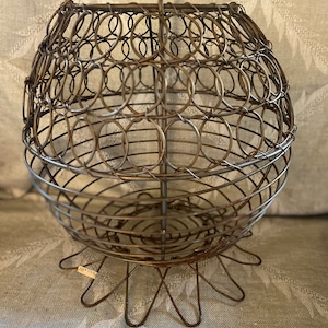 Può includere: Un cesto di frutta vintage in filo metallico, di forma rotonda, con un piedistallo a sei gambe. Il cesto è realizzato in filo metallico e ha una finitura marrone rustica. L'etichetta sul piedistallo recita "Country Home".