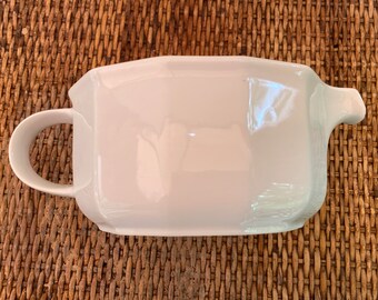 May include: White ceramic gravy boat with a handle and a spout. The boat is on a woven surface.
