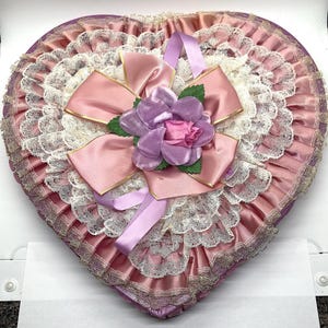 May include: A pink heart-shaped box with a lace trim and a pink satin bow with a purple flower.