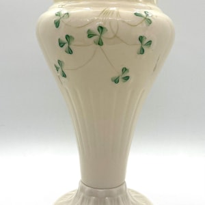 Puede incluir: Un jarrón de cerámica blanca con un borde festoneado y diseños de trébol verde. El jarrón tiene una base acampanada y un cuello delgado.