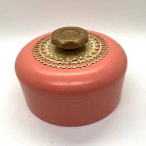 Könnte beinhalten: Eine vintage runde Schatulle in Rosa mit einem goldenen Filigranmuster und einem goldenen Knopf auf dem Deckel.