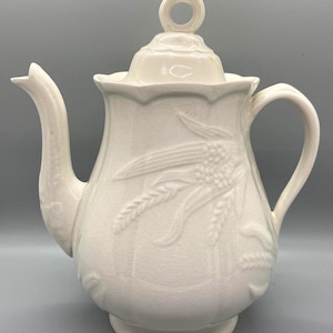Peut inclure: Théière en céramique blanche avec un couvercle et une poignée. La théière présente un motif en relief de tiges de blé et de fleurs.