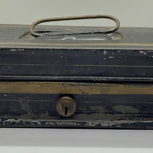 May include: A vintage black metal cash box with a brass-colored stripe and handle. The box has a keyhole and shows signs of wear, suggesting age. The top and sides have a decorative gold trim.